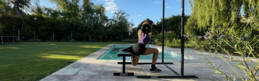 Mujer haciendo sentadillas búlgaras con banco plano SVAP y rack en casa